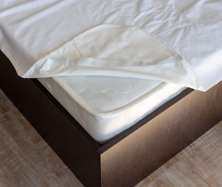 Corner of a wooden bed with a spring mattress and a waterproof mattress topper. Care of home furniture.